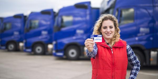 Truck driver proudly holding commercial driving license. In background you can see parked semi trucks.